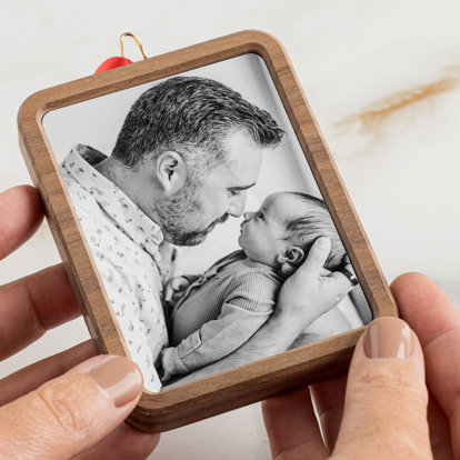 Hands holding a framed photo ornament of a dad nuzzling noses with his newborn baby. 