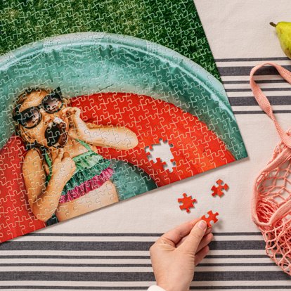 A hand finishing a custom photo puzzle on a table featuring an image of a young girl in a swimming pool eating an ice cream bar.
