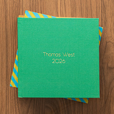 A stack of custom fabric photo books each with a different color cover. The book at the top of the stack has a bright green custom fabric cover and the text 'Thomas West 2026' in gold debossing. The second book has a diagonal stripe pattern with yellow and blue. And the book at the bottom has a mustard yellow fabric cover.