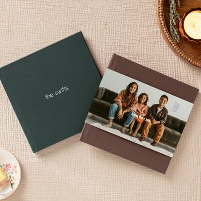 A closed photo book with a dark brown linen cover and photo dust jacket laying on top of a closed photo album with a dark green leather cover and silver foil stamped text.