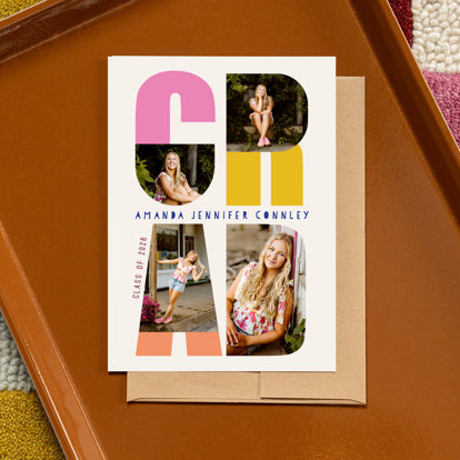 A graduation announcement set on a brown tray. The card features photos of a 2026 graduating senior inside the big block letters of ‘GRAD.’