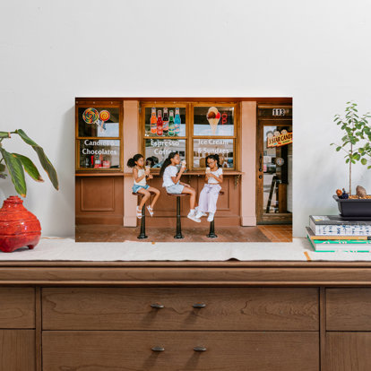 A large metal print displayed on a side table featuring a photo of three sisters sitting on stools outside a candy shop eating ice cream.