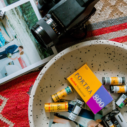 Kodak Portra 800 film box and assorted film rolls scattered in a speckled bowl beside a Contax film camera and printed photographs.