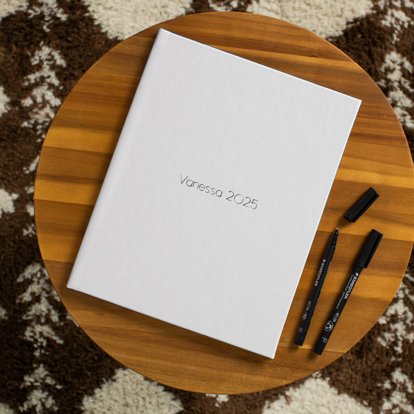 A closed photo guest book with the title 'Vanessa 2025' on the front cover and two black pens sitting next to it. 