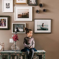 A young girl sitting on a bench below a gallery wall of framed family photos