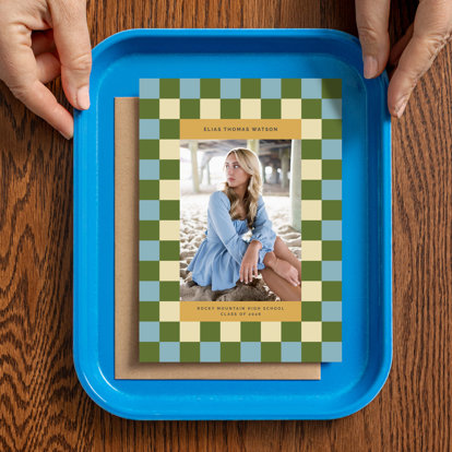 Hands holding a bright blue tray with a graduation announcement card set on it. The card features a 2026 graduating senior sitting on a beach under a pier with a  checkered border.