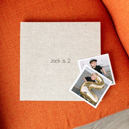 A photo book with a sand color linen cover and the title 'Jack is 2' sitting on an orange couch. Two photo prints of the birthday boy lay on the edge of the book. 