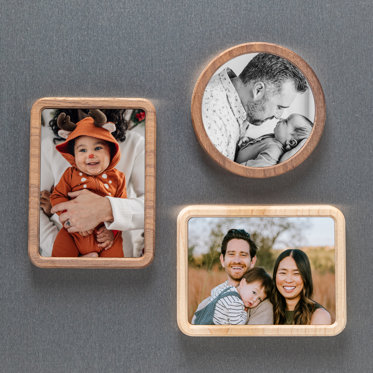 Three framed magnets displayed on a fridge. One is circular, the other two are rectangular shaped. The circle magnet features a family photo of mom and dad holding their toddler boy. One rectangle framed magnet features a baby in a reindeer costume. The other is of mother and father holding their young son.