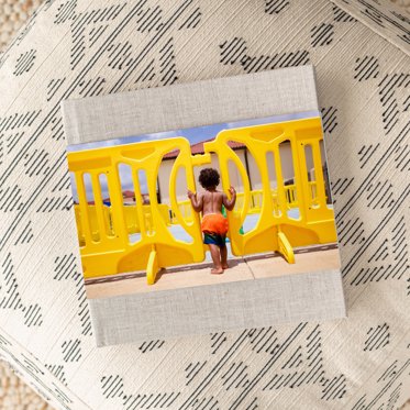 A premium hardcover photo book from Mpix on a table with a sand linen cover and a skinny dust jacket with a colorful photo of a toddler at play at the pool.