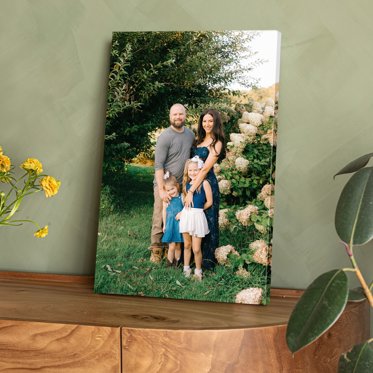 A canvas print displayed on a wood table featuring a couple and their two young daughters standing outside in front of a large hydrangea plant.