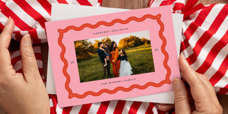 A close up of two hands holding a Christmas photo card with a pink background, a family photo, and personalized text around the photo that reads "Happiest Holidays 2025 The Murphy Family"