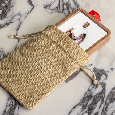 A rectangular shaped framed photo ornament half out of a brown woven bag set on a counter. The ornament features a dad with his two daughters. A red ribbon is attached to the ornament for hanging.