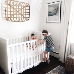 Two children at a crib beside a framed print on a white wall