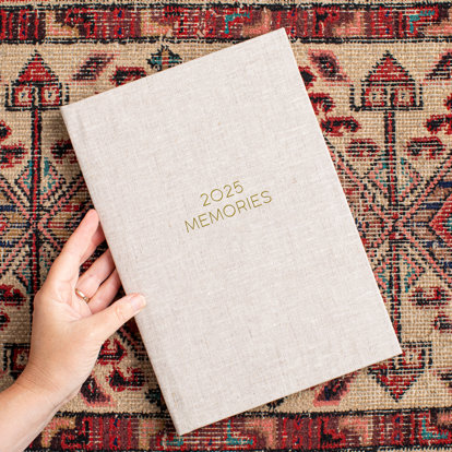 A closed 8x12 photo book with a sand color linen cover lying on a colorful cloth. The front cover has the text '2025 Memories.'