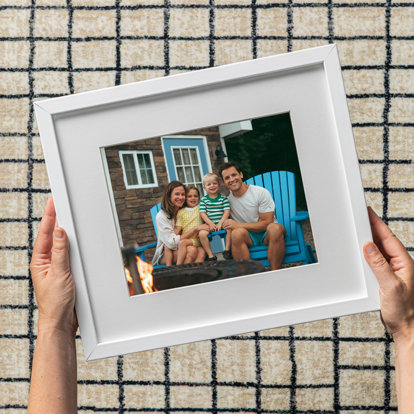 A framed print featuring a couple and their two young children sitting on blue adirondack chairs by a firepit. The photo has a white window mat and white metal frame.