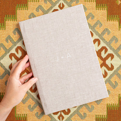 A closed premium hardcover photo book with a sand color linen cover and the text "J + A" in silver debossing. 