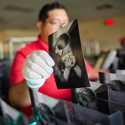 An Mpix photo lab worker sorting prints. The 5x7 black and white print he's holding features a toddler girl laughing.