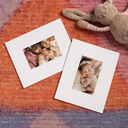 Two 4x6 matted prints with newborn imagery laid out on a rug next to a stuffed animal.