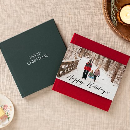 A closed photo book with a red linen cover and photo dust jacket laying on top of a closed photo album with a dark green leather cover and silver foil stamped text that reads "MERRY CHRISTMAS".