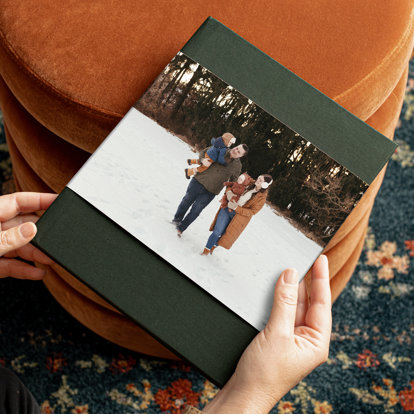 Hands holding a closed photo book with an evergreen linen cover and a skinny dustjacket featuring a family of four walking in the snow.