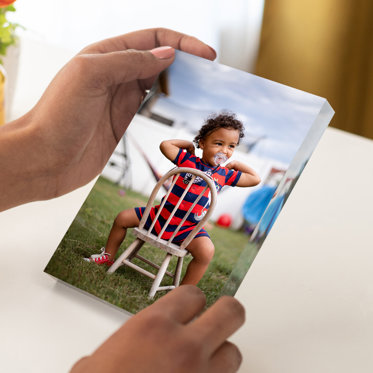 Hands holding an acrylic photo block featuring a toddler sitting backwards on a chair and smiling with a pacifier in his mouth. 