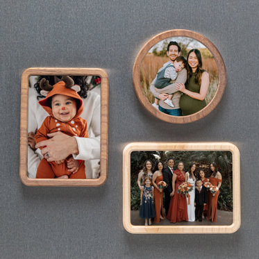 Three framed photo magnets displayed on a fridge. One is circular, the other two are rectangular shaped. The circle magnet features a family photo of mom and dad holding their toddler boy. One rectangle framed magnet features a baby in a reindeer costume. The other is of a family picture at a wedding.