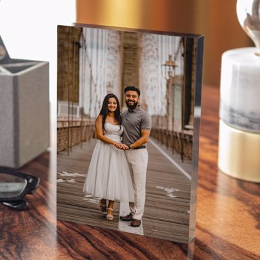 An Acrylic Photo Block from Mpix sitting on a desk next to a pair of glasses & featuring engagement photos of a couple standing on a bridge.
