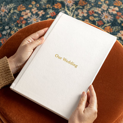 Hands holding an Mpix signature photo album with a white leather cover and 'Our Wedding' in gold debossing on the front cover.