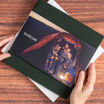 A close up of hands holding a closed hardcover photo book with a dark green linen cover and dust jacket featuring an image of a family camping in a tent.