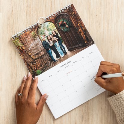 Hands filling out the date grid of a calendar that also includes spiral binding on top and a family photo above the grid.