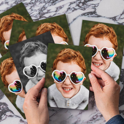 Hands holding a close-up photo print of a young red-haired boy wearing heart-shaped sunglasses.